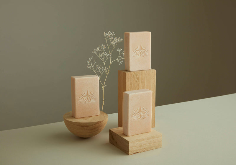 Three pale pink citrus rose salt bars sitting on wooden blocks with delicate dried white flowers in the background. Our logo is seen clearly on the front of the salt bars